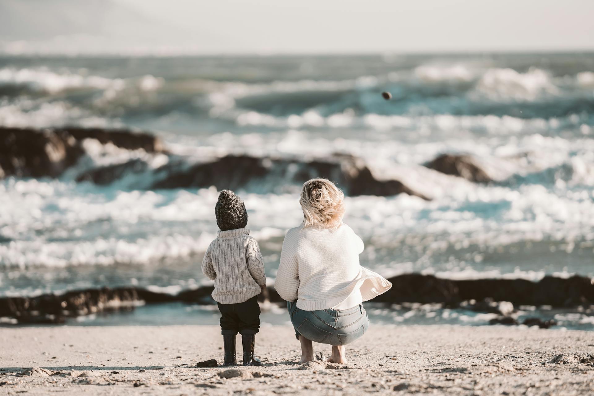 A mother and child share quality time on a serene beach, facing the ocean waves.