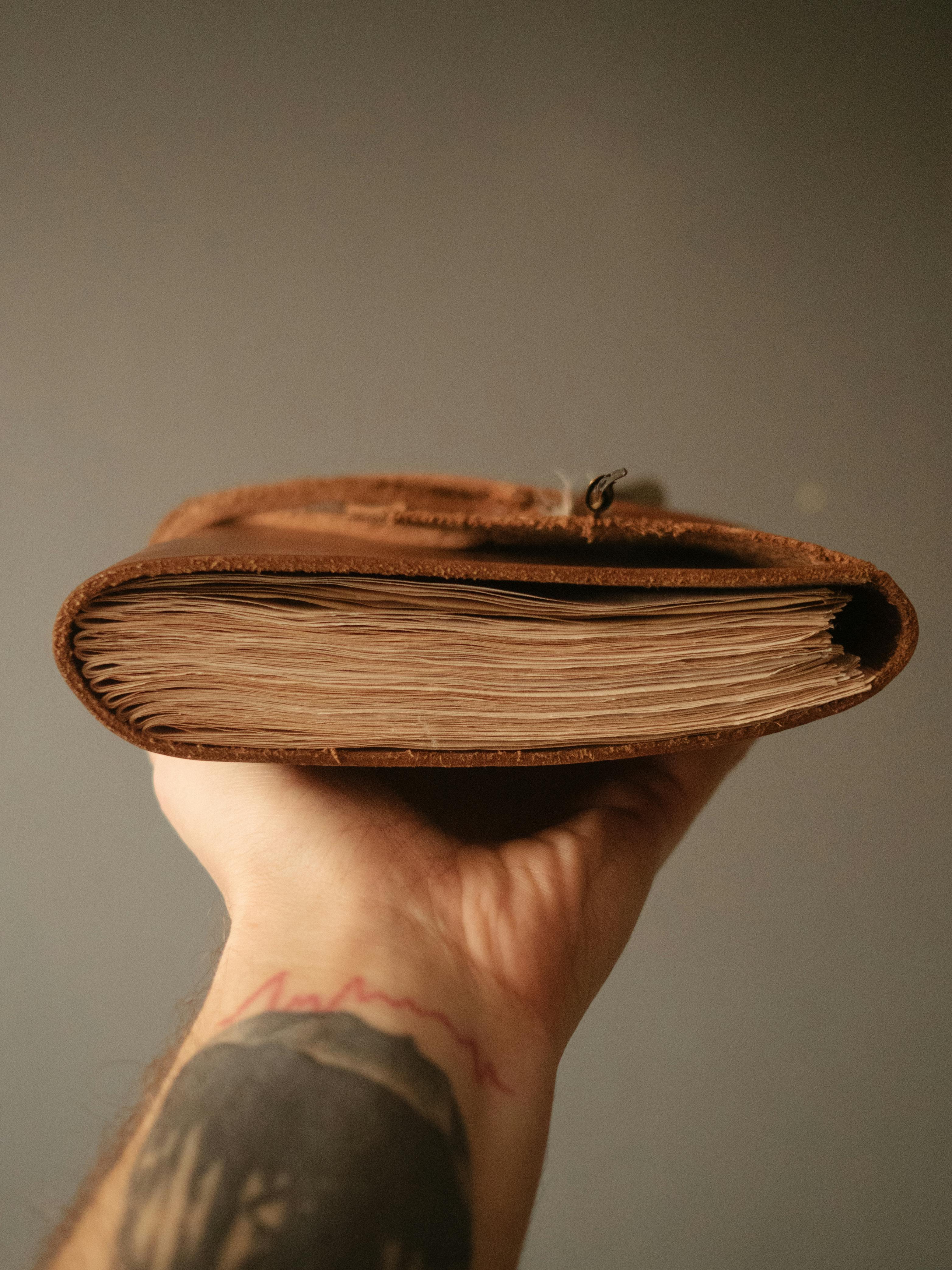 Un primer plano de una mano tatuada sosteniendo un diario de cuero rústico contra un fondo neutro.