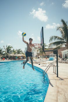 A man performs a basketball dunk near a swimming pool during a sunny vacation.