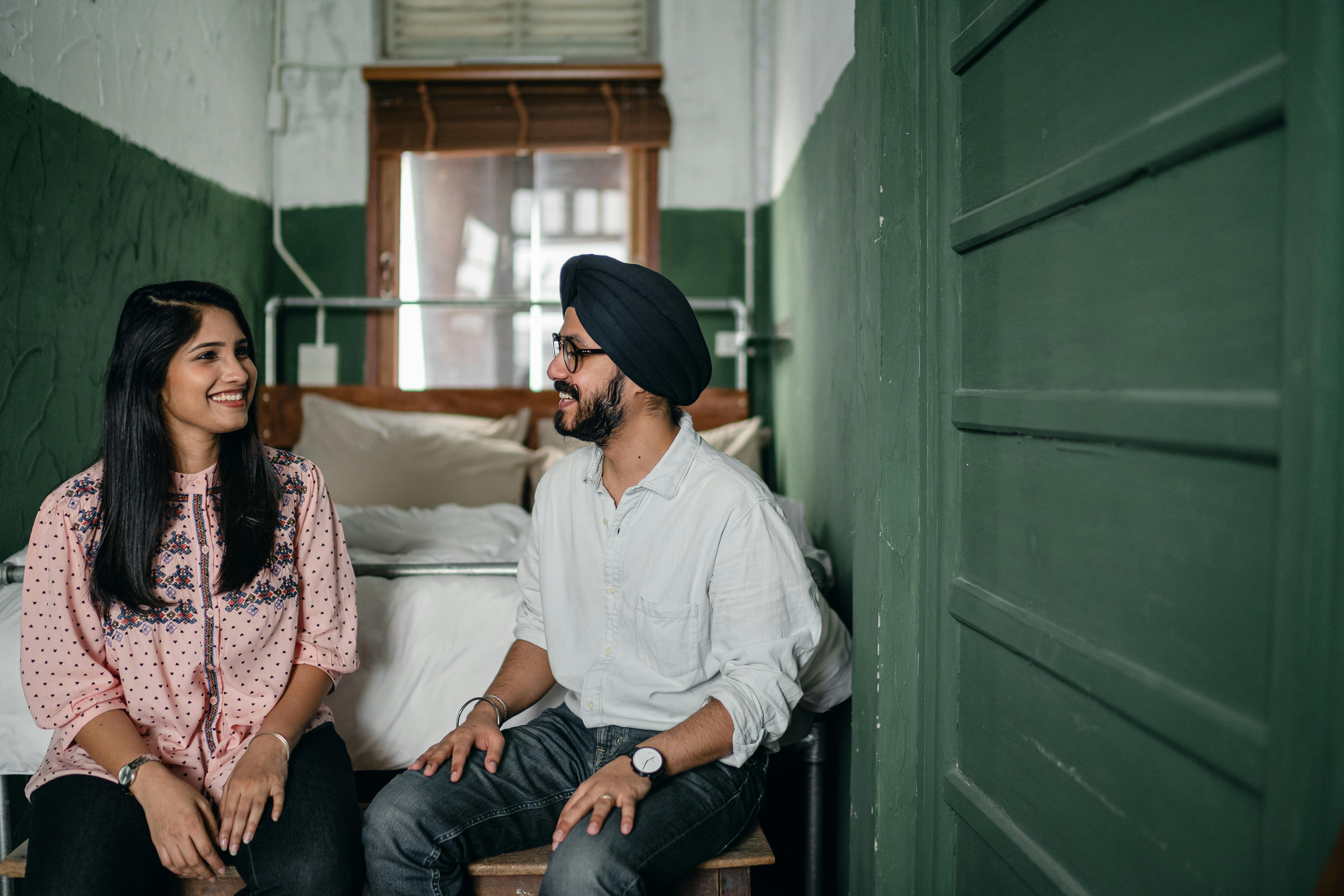 Smiling Indian couple in casual clothes and turban sitting on bench against bed in narrow green bedroom and chatting while looking at each other happily