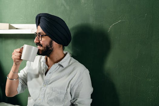 A cheerful man wearing a turban enjoys a cup of tea indoors with a green wall backdrop.