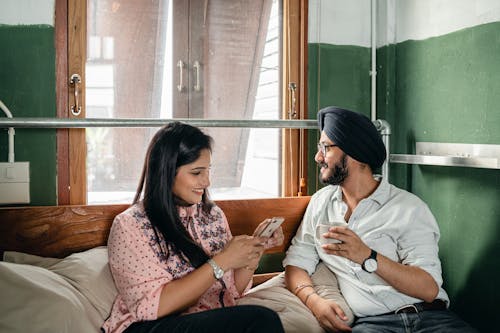 Optimistic smiling excited Indian couple in casual clothes and turban using mobile phone and chatting while relaxing on comfortable bed in sunny room