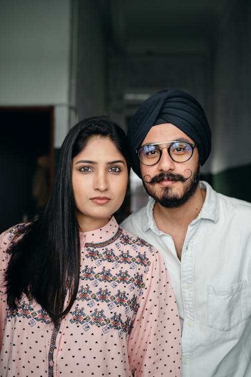 Portrait of positive young Sikh couple in casual outfits wearing turban and eyeglasses standing close to each other and looking at camera