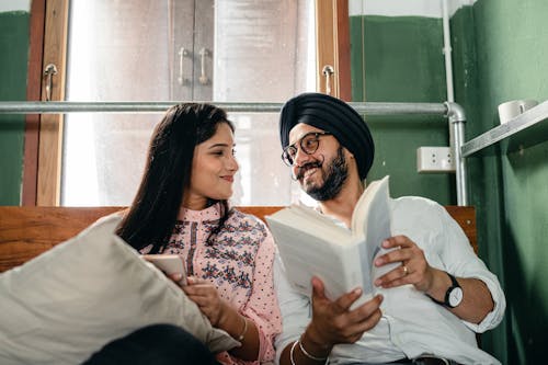Loving young Indian couple in casual wear and turban with book and mobile phone resting on comfy bed and looking at each other happily