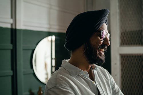 Side view of bearded positive laughing Indian male in eyeglasses wearing casual shirt and turban standing near green and white wall and mirror and looking away happily