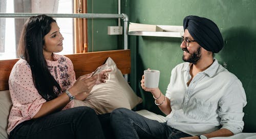 Free Happy optimistic Sikh couple in casual clothes and turban with smartphone and cup of hot drink relaxing on comfortable bed while smiling and looking at each other in daylight Stock Photo