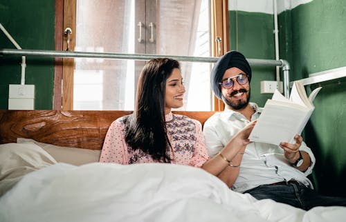 Happy smiling young Sikh couple in turban and casual clothing reading interesting book while resting together on cozy bed in narrow green room near window in daylight