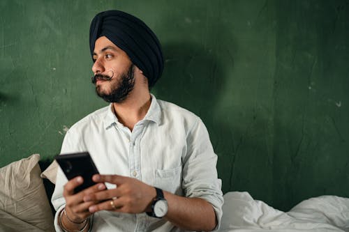 Serious bearded Indian male wearing turban sitting on bed against green old wall while browsing mobile phone and looking away pensively