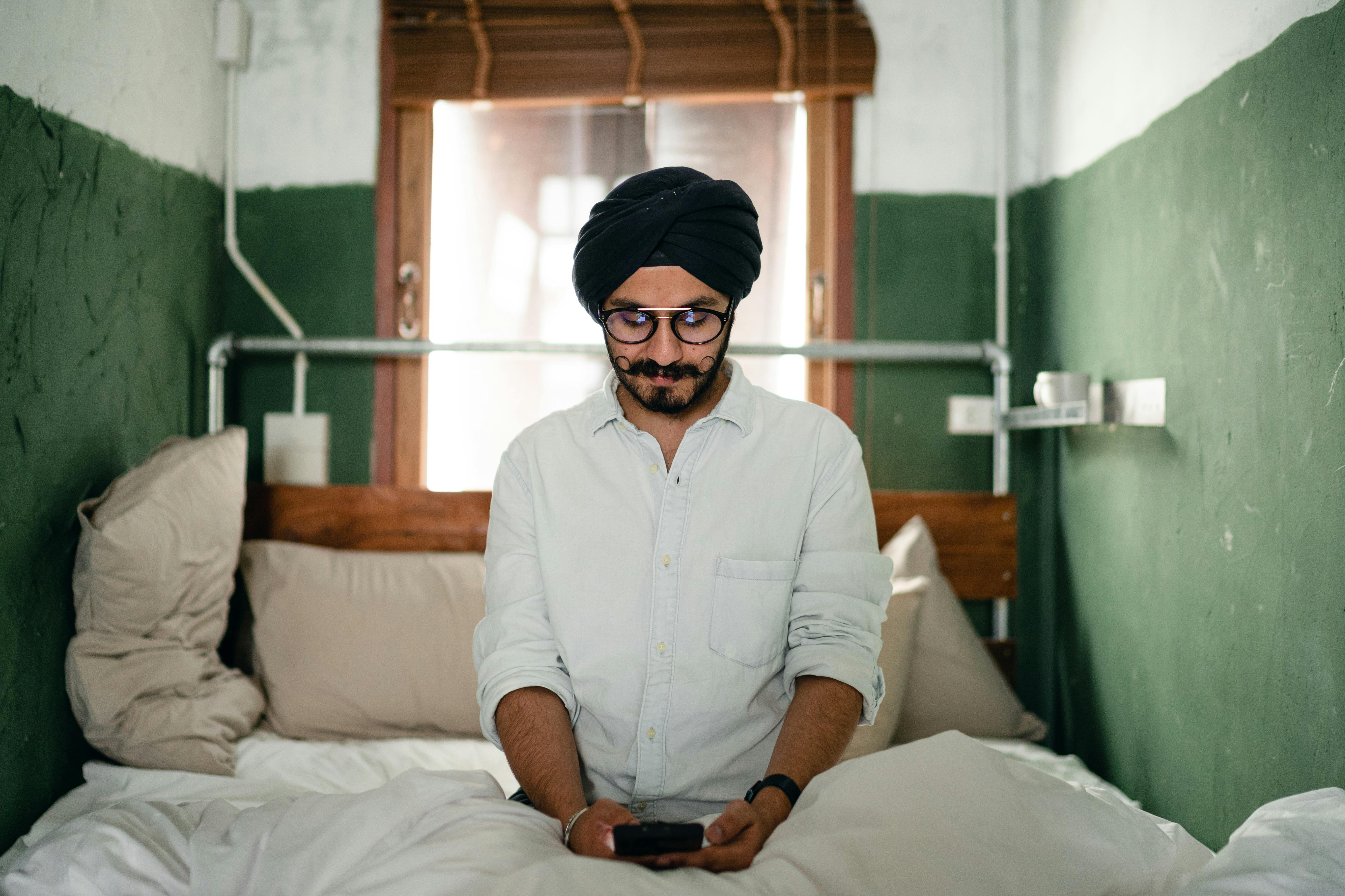 pensive man in turban using smartphone on bed