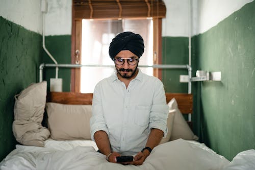 Pensive man in turban using smartphone on bed