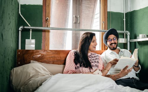 Happy Indian couple of husband in turban and glasses with handlebar mustache and good looking wife reading book and laughing while resting on big wooden bed