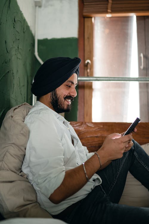 Side view of young Indian male in casual clothes and turban with handlebars sitting on cozy bed and messaging on cellphone