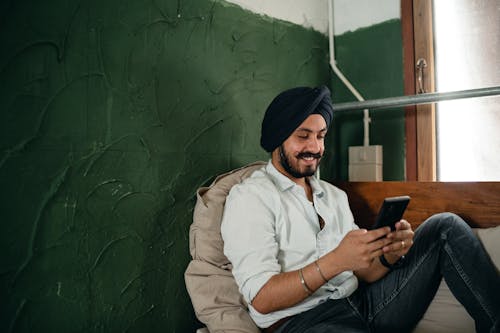 Free Smiling man in turban using smartphone while relaxing at home Stock Photo