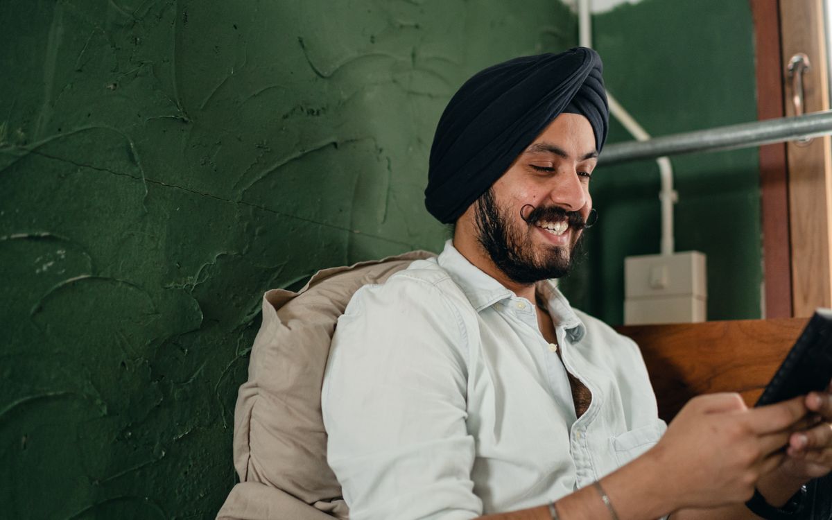 Young bearded Indian man in white shirt and dark turban leaning on pillow and browsing net on smartphone while spending time at home