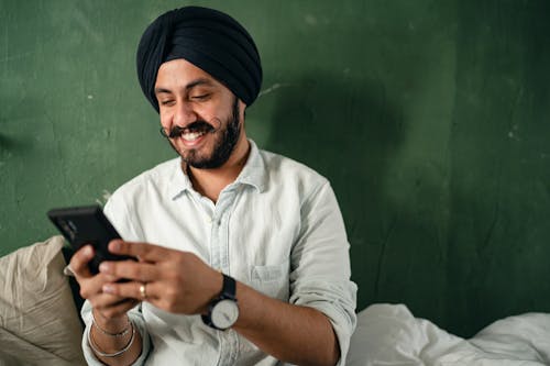 Free Young Indian male with black beard and mustache in casual shirt and turban messaging on smartphone and smiling happily while sitting on bed near green wall at home Stock Photo