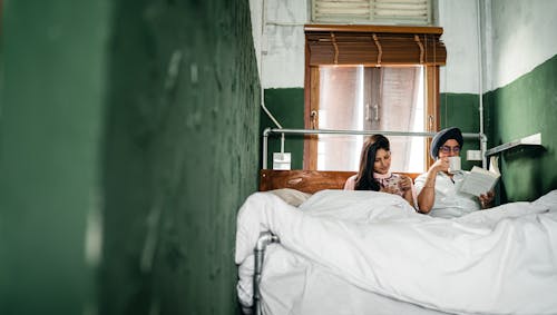 Indian couple of wife scrolling through news feed on smartphone and husband enjoying coffee and reading book on wooden bed in room with roughly plastered green walls