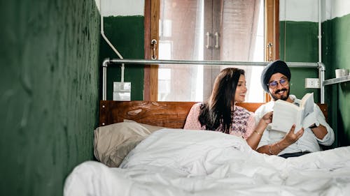 Free Young Indian husband and wife talking and looking through interesting book while relaxing on wide wooden bed in room with plastered walls Stock Photo