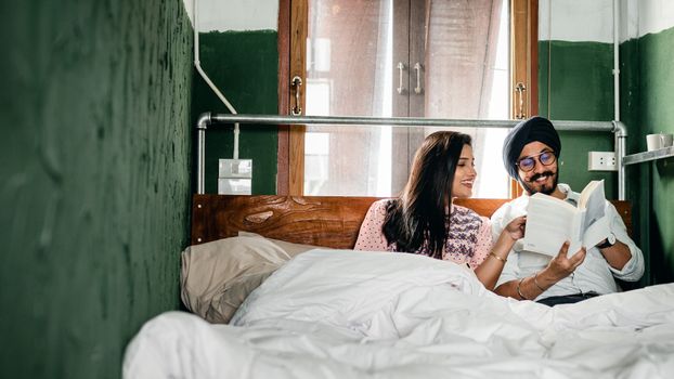 Young Indian husband and wife talking and looking through interesting book while relaxing on wide wooden bed in room with plastered walls