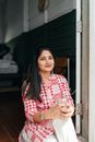 Happy young Indian female in long red and white checkered shirt and white pants sitting on threshold of house with glass of water and looking away on sunny day