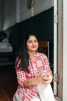 Happy young Indian female in long red and white checkered shirt and white pants sitting on threshold of house with glass of water and looking away on sunny day