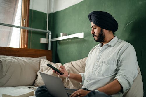 Bearded Indian businessman in casual clothes with serious expression on face texting on mobile and surfing net on laptop while working in comfort of quiet room