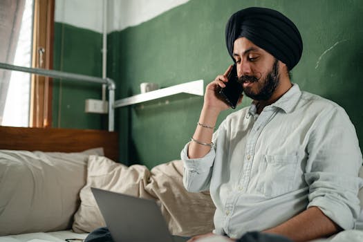 South Asian man sitting on a bed using a smartphone and laptop, showing modern remote work lifestyle.