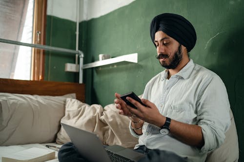 Free Concentrated bearded Sikh male wearing turban sitting on bed with pillows in green room near window with netbook and browsing smartphone pensively Stock Photo