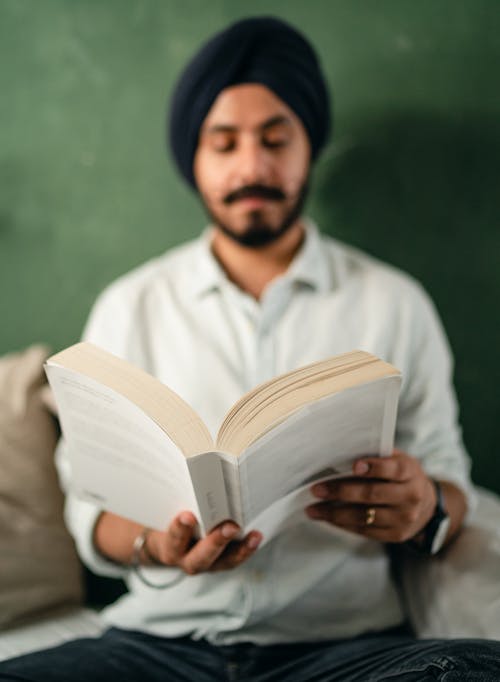 Focused bearded Indian male in casual shirt and black turban sitting against green wall and reading interesting book