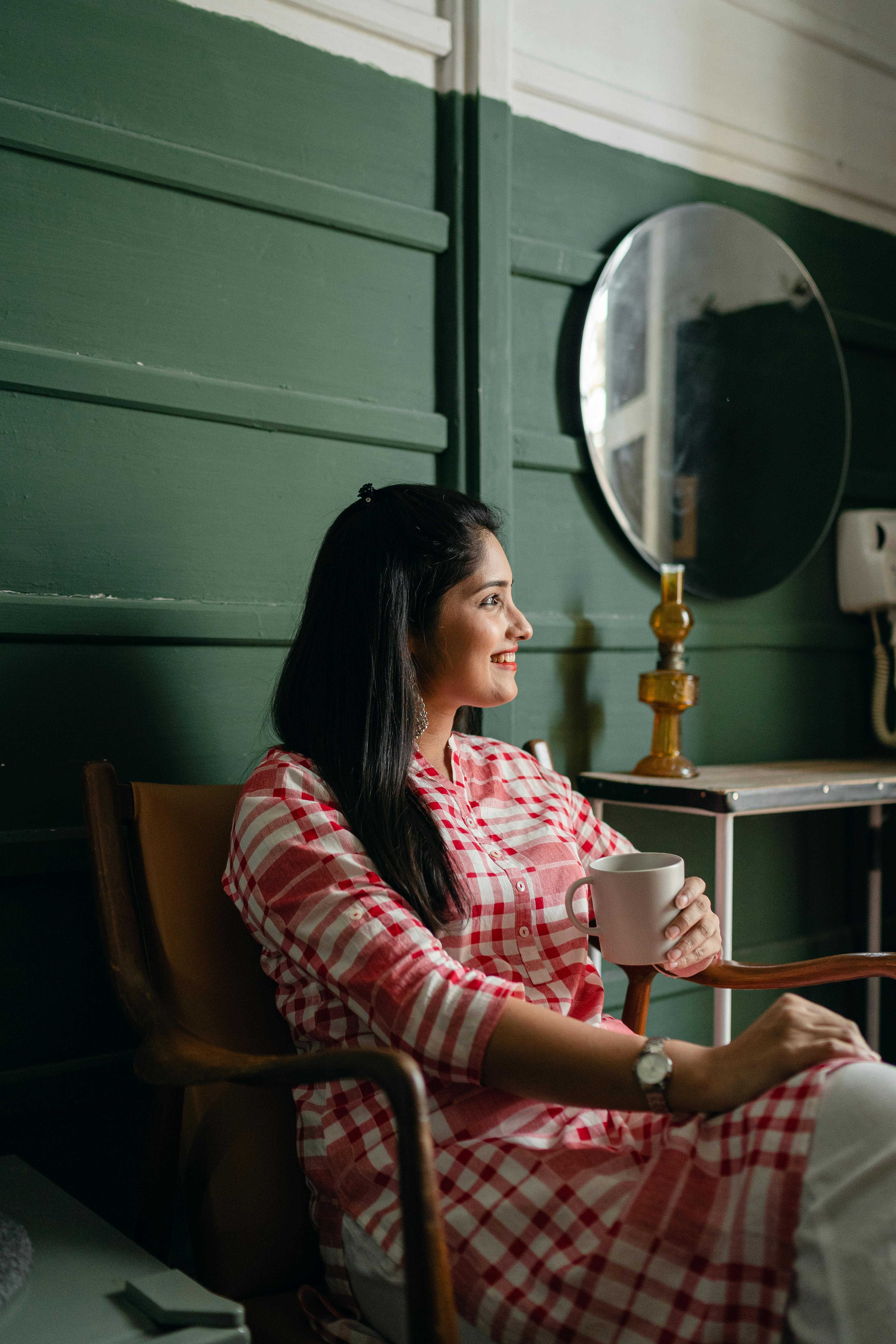 Side view of young positive Indian lady in trendy traditional clothes resting on chair with cup of drink against green wall near mirror