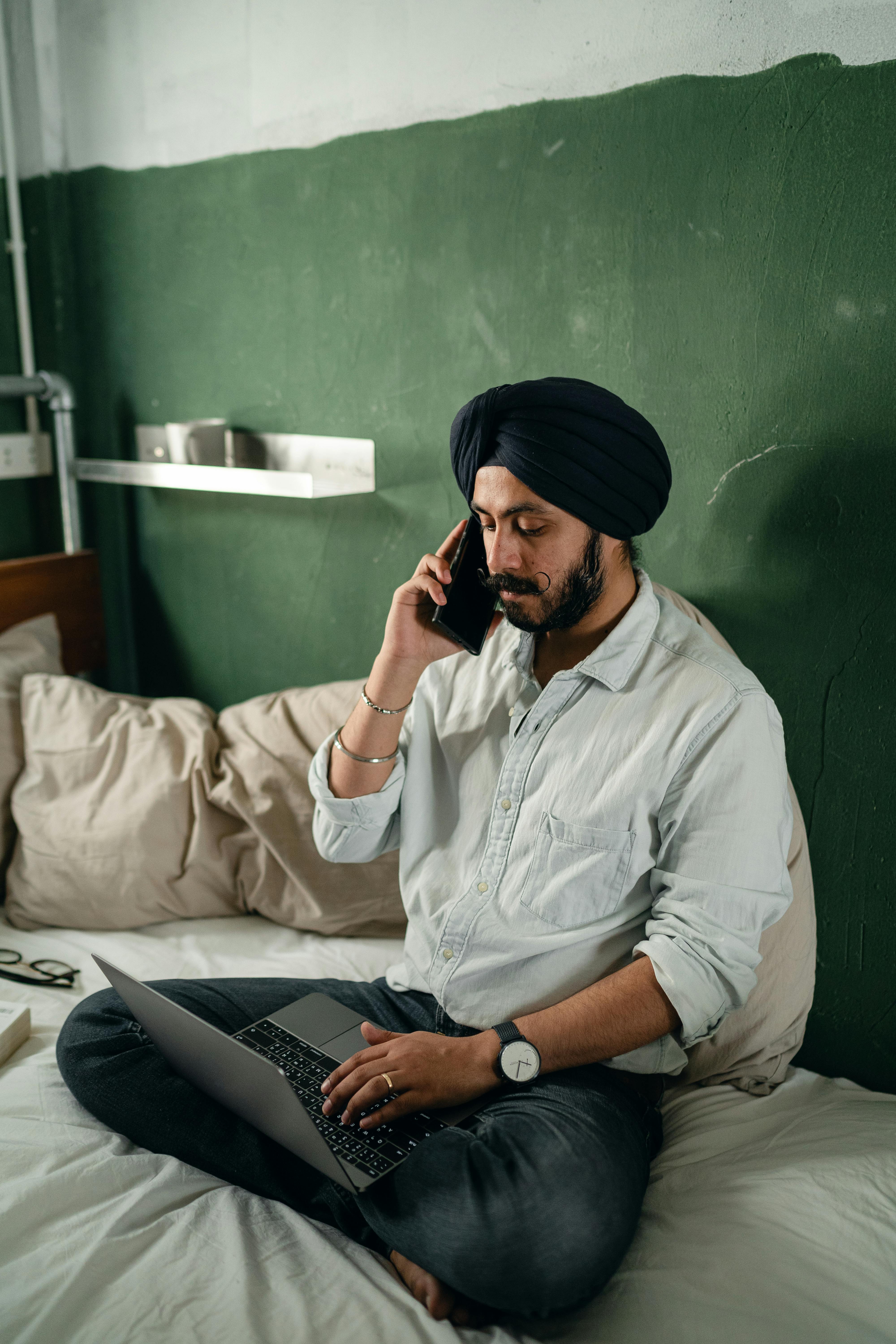 Focused bearded Indian male freelancer wearing turban sitting on bed against green wall in old room and having conversation via cellphone while working on netbook remotely