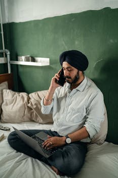 Focused bearded Indian male freelancer wearing turban sitting on bed against green wall in old room and having conversation via cellphone while working on netbook remotely