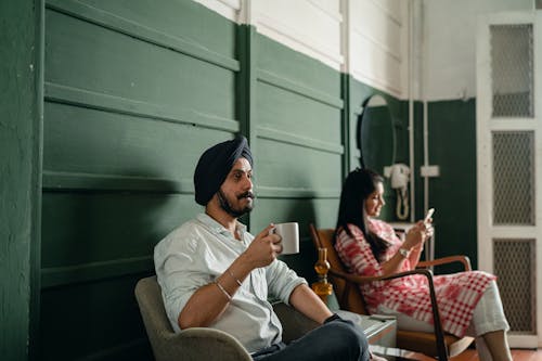 Side view of thoughtful Sikh male in turban with cup of hot beverage sitting on chairs with Indian woman in traditional pink wear with smartphone in room with green walls