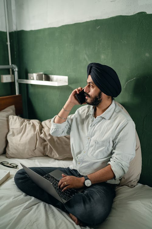 Full length of focused bearded man sitting on bed against green wall while working on netbook and having conversation via smartphone