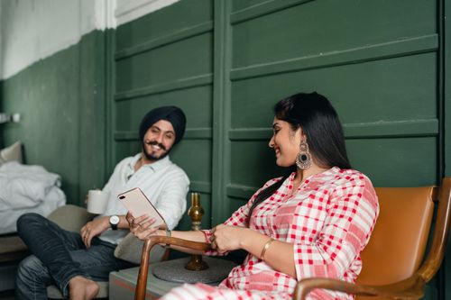 Free Happy young Indian couple in stylish traditional clothes sitting on leather chairs and browsing cellphone in bedroom with green walls Stock Photo