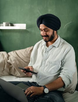 Cheerful bearded Indian male wearing turban sitting with legs crossed on comfortable bed with laptop on laps and browsing smartphone