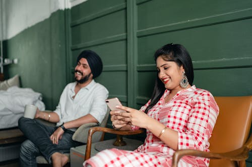 Positive young Sikh couple in casual traditional clothes resting on leather chairs against green wall while browsing mobile phone and laughing