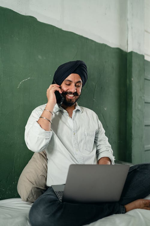 Cheerful man speaking on smartphone and working on laptop