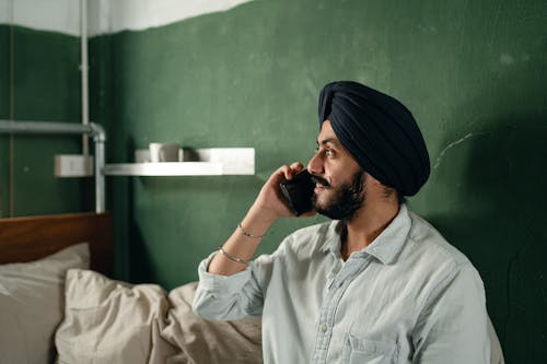 Side view of handsome Indian male engaged in pleasant phone conversation while sitting in simply furnished room and looking away smiling softly