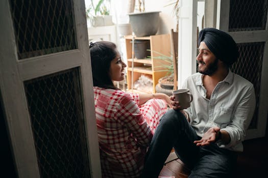 Positive young Indian spouses having lively conversation and drinking coffee while relaxing on threshold of sunlit enclosed balcony