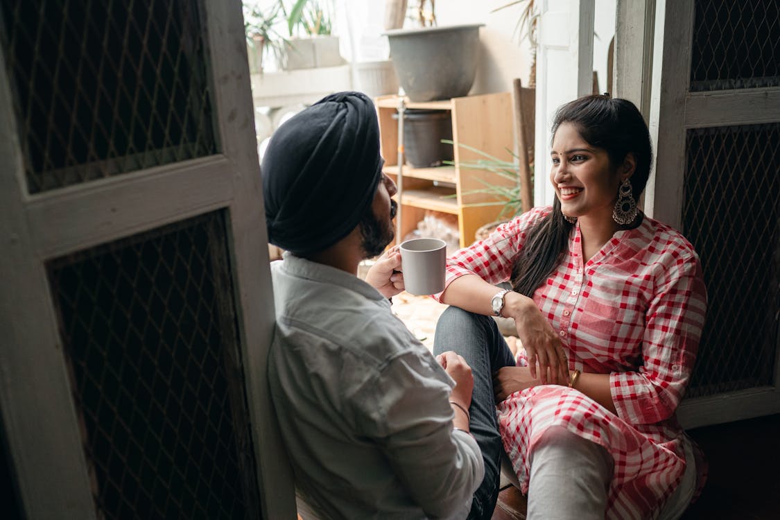 Free Cheerful Indian couple of smiling lady listening to boyfriend telling amusing story and drinking coffee on floor at threshold of loggia on sunny day off Stock Photo