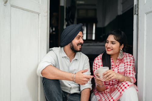 Elegant Indian wife with cup of coffee listening to handsome Indian husband while sitting at door of house