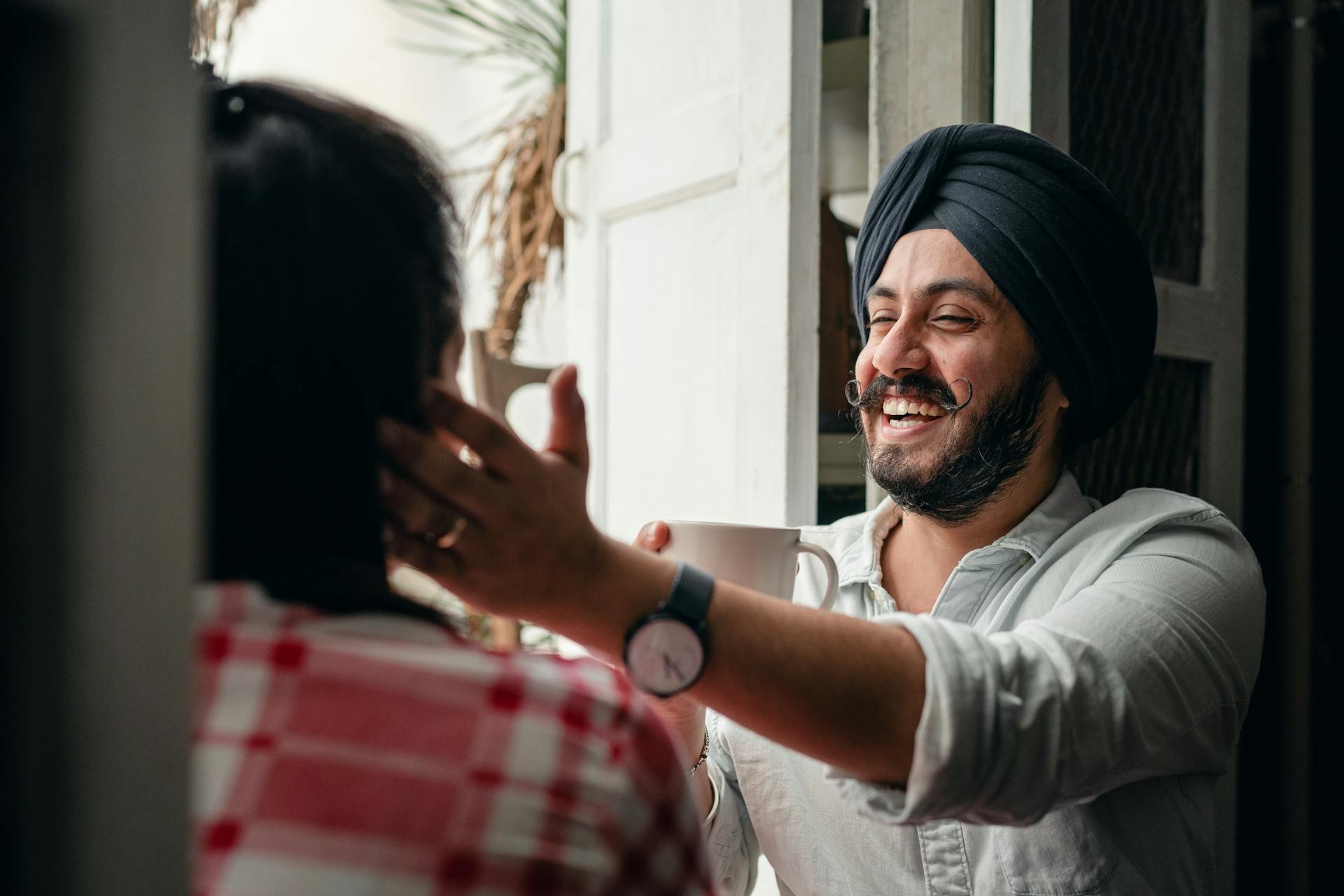 Happy modern Indian man with cup of coffee laughing heartily and touching cheek of beloved woman while enjoying time at home