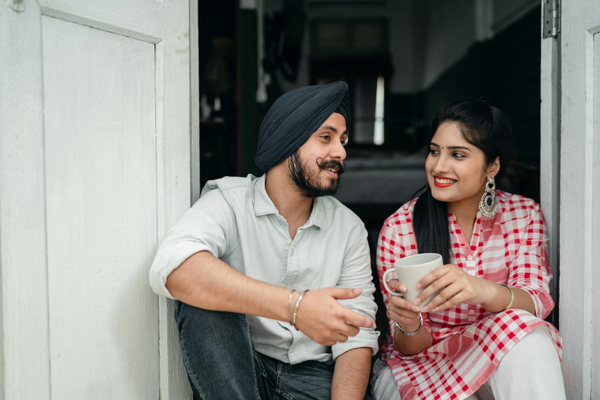 Positive Indian spouses in casual outfits sharing interesting stories while drinking morning coffee on doorstep of house