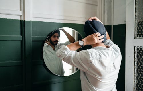 Confident bearded Sikh male wearing denim shirt adjusting traditional headwear standing in front of mirror on wall at home while taking care of appearance