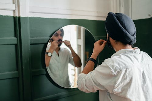 Stylish bearded Sikh male wearing denim shirt and turban making mustache hairdo standing in front of mirror on wall at home while taking care of appearance
