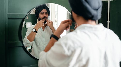 Back view of young Indian male with twisted mustache wearing denim shirt and turban taking care of beard standing against mirror on wall during daytime