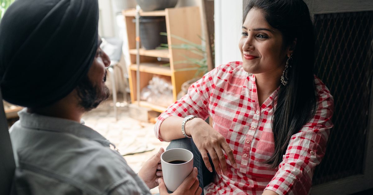 Cheerful young wife with bindi on forehead wearing plaid dress sitting in doorway against street kitchen with young bearded husband in turban while communicating and drinking tea and looking at each other