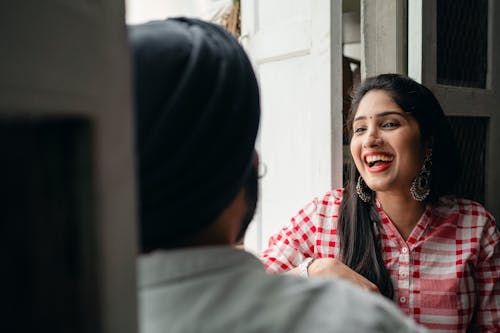 Excited young attractive female with bindi on forehead wearing large earrings with authentic design and checkered cloth sitting in doorway with blurred husband wearing turban during free time