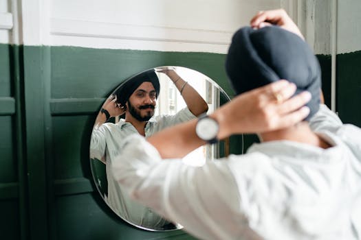 Back view of satisfied young Indian male hipster with twisted mustache wearing denim shirt and turban standing against mirror on wall while taking care about appearance