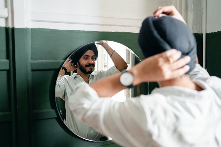 Mirror Reflection Of Stylish Sikh Man Adjusting Black Turban At Home